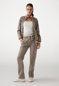 Brauner gemusterter Trainingsanzug mit weißen Blumenmuster, bestehend aus einer zip-up Jacke und Hose mit Kordelzug, kombiniert mit weißen Turnschuhen.