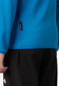 Camicia a maniche lunghe blu realizzata in un tessuto liscio, con etichetta Polartec e un capo nero a contrasto nella parte inferiore.