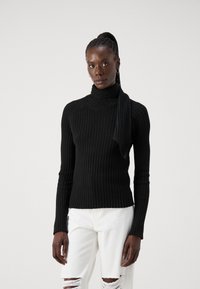 Must mustriba turtleneck-sweater koos kinnitatud salliga, millel on keha lähedane siluett ja pikad varrukad. Kombineeritud kulunud valgete teksadega.