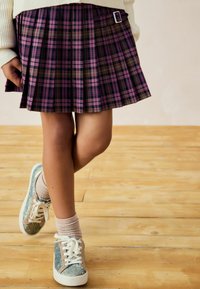 Lila und schwarze karierte Faltenrock mit einer Schnallendetail, kombiniert mit glitzernden Sneakers und Kniestrümpfen. Hintergrund aus Holzfußboden.