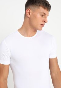 Fehér, rövid ujjú póló kerek nyakkivágással, sima, rugalmas anyagból készült. Szoros illeszkedésű és minimális varrásokkal rendelkezik.