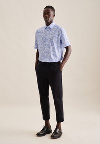 Camicia a maniche corte blu e bianca con motivo paisley abbinata a pantaloni neri a gamba affusolata e scarpe nere con lacci. Colletto semplice e orli dei pantaloni risvoltati.
