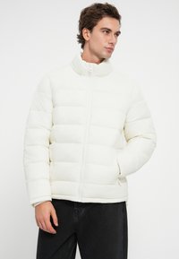 Φουσκωτό μπουφάν σε εκρού, με επένδυση καπιτονέ, ψηλό γιακά, εμπρός φερμουάρ και πλαϊνές τσέπες με φερμουάρ. Μαλακή υφή.