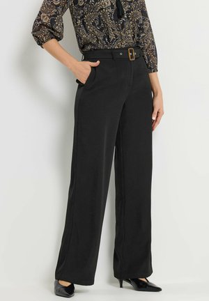 Vrouw draagt zwarte wijde broek met hoge taille en bijpassende riem, paisley blouse en zwarte puntige hakken, hand in zak.