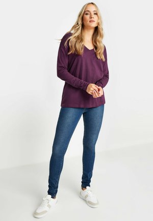 Lila Langarm-V-Ausschnitt-Oberteil kombiniert mit blauen Skinny-Jeans und weißen Sneakern mit beigen Akzenten; glatte Textur, lässige Passform.