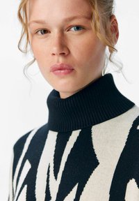 Pull en col roulé tricoté en noir et crème avec des motifs zigzag abstraits ; col à côtes et texture douce. Gros plan sur le visage et le vêtement.