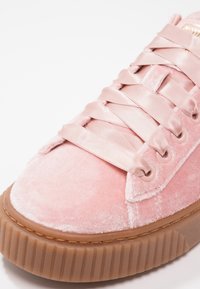 Gros plan sur une chaussure de sport en velours rose avec de larges lacets en satin et une semelle épaisse en caoutchouc marron aux bords rainurés.