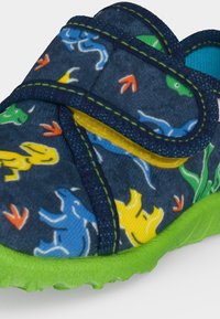 Gros plan sur une chaussure d’enfant bleue avec des motifs de dinosaures colorés, des empreintes orange, ainsi qu’une semelle et un liseré vert vif. Bride Velcro visible.