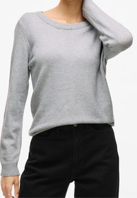 Personne portant un pull à manches longues gris clair et un pantalon noir taille haute, avec un bras levé et la main touchant la nuque.