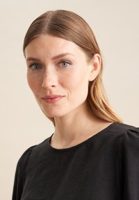 Top in lino nero con scollatura rotonda e morbidi arricciamenti sulle spalle, indossato da una donna con capelli lunghi e lisci, su uno sfondo neutro.