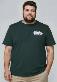 Tmavě zelené bavlněné tričko s bílou grafikou, na níž je nápis "BETONOVÁ JUNGLE" a design siluety města na hrudi.