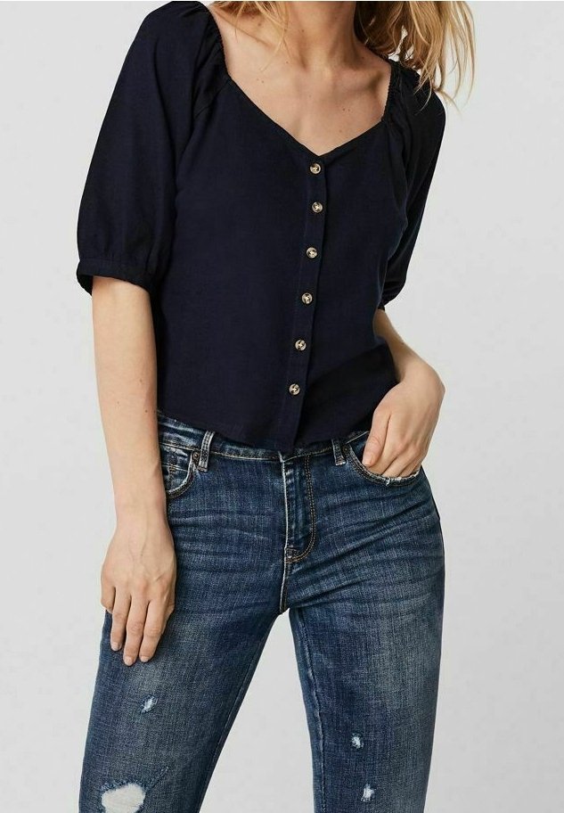 Femme portant un chemisier noir boutonné à manches bouffantes et un jean bleu usé, une main dans la poche, sur fond uni.