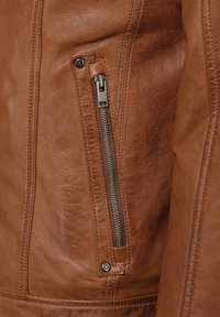 Braune Lederjacke mit einer Seiten tasche, die mit einem silbernen Reißverschluss und einem Druckknopfverschluss versehen ist, strukturierter Oberfläche und Nahtdetails.