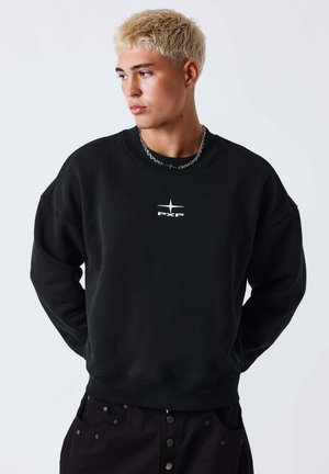 Jeune homme avec des cheveux blonds courts portant un sweat-shirt noir avec un logo étoile blanc et un collier chaîne, regardant vers sa gauche les mains derrière le dos.