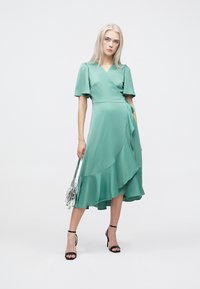 Mētas zaļas krāsas wrap kleita ar īsām uzpūstām piedurknēm un plūstošu rievotu apakšmali. Valkāta ar melnām siksniņām apaviem un tur rožaini caurspīdīgu somiņu.