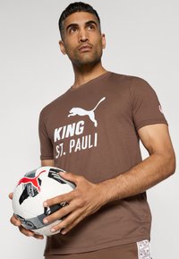 Brun kortærmet t-shirt med hvid "KING ST. PAULI" tekst og logo, parret med en mønstret sort og hvid fodbold med et rødt logo.