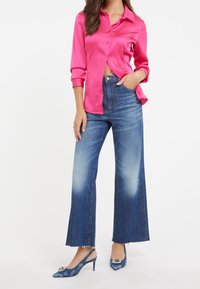 Chemise en satin rose vif à manches longues, jeans en denim bleu taille haute à jambes larges, et chaussures à talons hauts bleu à motif avec un détail décoratif.