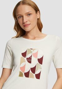 Weißes T-Shirt mit kurzen Ärmeln, das einen geometrischen Mehrfarben-Druck in Bordeauxrot, Pink, Beige und goldfarbenen Akzenten zeigt, mit überlagertem Text.