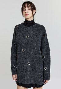 Maglioncino grigio oversize con dettagli in occhio di pernice argentati, polsini a coste e lavorazione a maglia testurizzata. Indossato sopra un dolcevita nero, con vestibilità comoda.