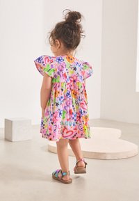 Vestito colorato con maniche corte a sbuffo, caratterizzato da una stampa vivace e giocosa di fiori, cuori e faccine sorridenti. Sandali beige completano il look.