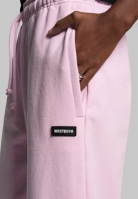Lys pink sweatpants lavet af blødt stof, med elastisk talje, sidelommer og et sort mærke med "WRSTBHVR" branding.