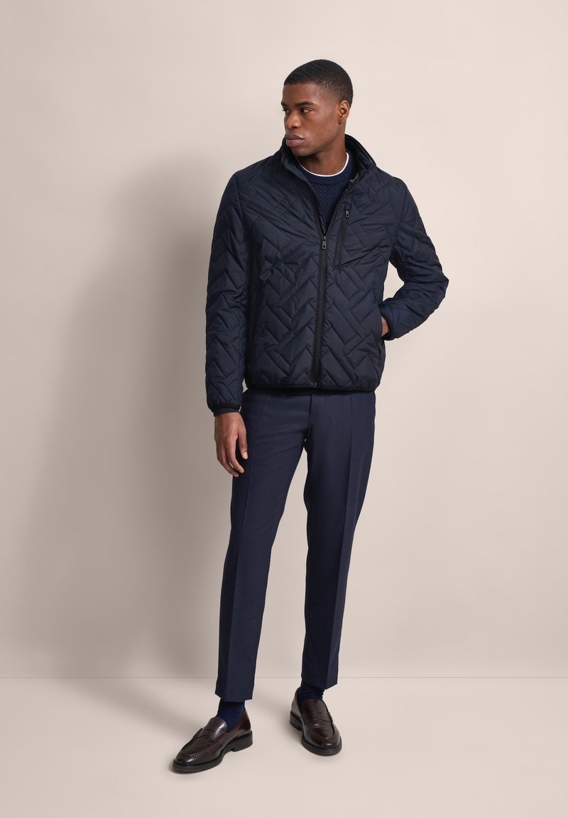 Marineblauer gesteppter Blazer mit Stehkragen, Reißverschluss und subtilem Zickzackmuster; kombiniert mit dunklen Hosen und braunen Slippern.
