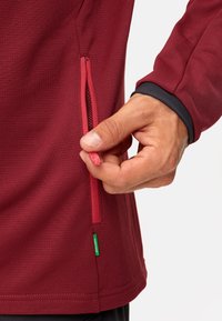Maglietta sportiva rossa in tessuto testurizzato, con tasca laterale con cerniera, dettagli neri ai polsini e etichetta verde del marchio all'orlo.