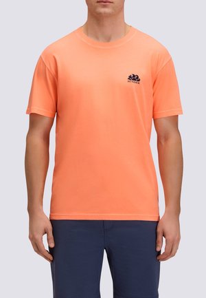 Uomo che indossa una maglietta Sundek arancione brillante a maniche corte e pantaloni blu scuro, in piedi davanti a uno sfondo neutro.