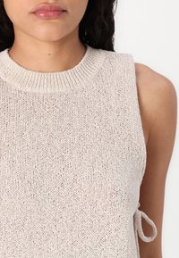 Débardeur en tricot beige clair sans manches avec un col rond côtelé et des détails de nœud sur le côté. La texture semble douce et respirante.