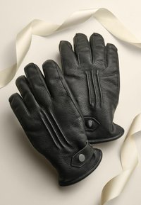 Zwarte leren handschoenen met een textuurafwerking, voorzien van geribbelde ontwerpaccenten en een drukknoopsluiting. Geplaatst naast een crèmekleurig lint.