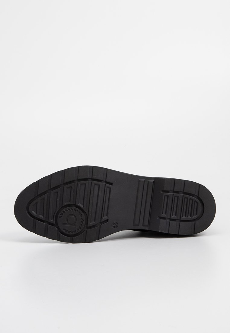Semelle de chaussure en caoutchouc noir présentant des rainures horizontales et verticales, un talon arrondi et un détail de logo circulaire près du centre.