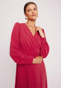 Sukienka kopertowa w głębokim różu, z długimi bufkami, dekoltem w kształcie litery V oraz detalami guzików przy mankietach. Gładki, lekki materiał.