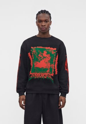 Jeune homme aux cheveux tressés portant un sweat-shirt noir avec un grand motif graphique abstrait rouge et vert et des motifs de flammes rouges sur les manches.