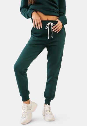 Donkergroene joggingbroek van zachte stof, met elastische boorden en een tailleband met trekkoord. Gecombineerd met witte sneakers.