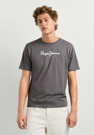 Joven vestido con una camiseta gris carbón Pepe Jeans London y pantalones blancos, de pie contra un fondo liso gris claro.