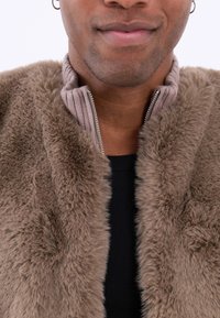 Jachetă din blană sintetică maro cu guler ribbed, închidere prin fermoar și textură moale. Articolul negru de îmbrăcăminte de dedesubt este vizibil în apropierea gulerului.