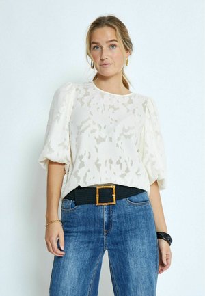 Weiße Bluse mit Puffärmeln und subtil strukturiertem Muster, kombiniert mit hochtaillierten blauen Jeans und einem schwarzen Gürtel mit einer goldenen quadratischen Schnalle.