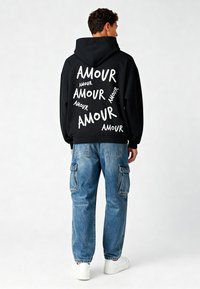 Felpa nera con il testo "AMOUR" bianco di varie dimensioni sulla schiena. Abbinata a jeans cargo blu chiaro e sneakers bianche.