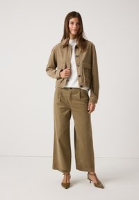 Braune Cordjacke mit Fronttaschen, Knopfverschluss und elastischem Saum, kombiniert mit weit geschnittenen braunen Cordhosen. Weißes Rundhalsshirt.