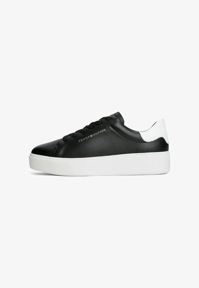 Schwarzer Low-Top-Sneaker mit weißer Ferse und dicker weißer Sohle, goldenem "Tommy Hilfiger"-Logo an der Seite und schwarzen Schnürsenkeln.