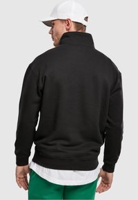Black sweatshirt with high collar, made of soft material. Features ribbed cuffs and hem. Worn over a white shirt and green pants.