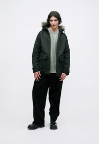Chaqueta acolchada negra con capucha forrada de piel, bolsillos frontales y cierre de cremallera, llevada sobre una camisa gris clara y pantalones anchos negros.