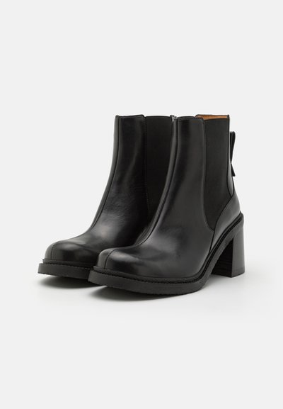 Botas de tobillo de cuero negro con puntera redondeada, paneles laterales elásticos y un tacón alto y grueso. La suela texturada contrasta con la parte superior lisa.