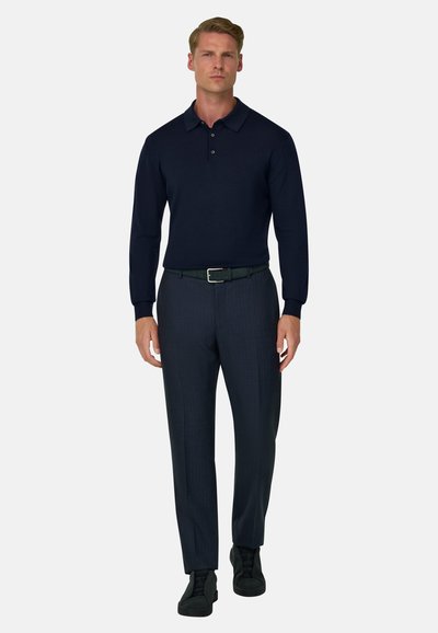 Homme marchant en avant portant un polo bleu marine foncé à manches longues, un pantalon foncé, une ceinture noire et des chaussures noires sur un fond blanc uni.