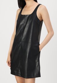 Kvinna i en ärmlös svart läderklänning med fyrkantig halsringning och händerna i sidfickorna mot en enkel bakgrund.