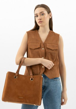 Borsa a tracolla in suede marrone con una forma strutturata, doppi manici superiori e dettagli in metallo dorato. Il modello indossa una blusa senza maniche marrone abbinata.