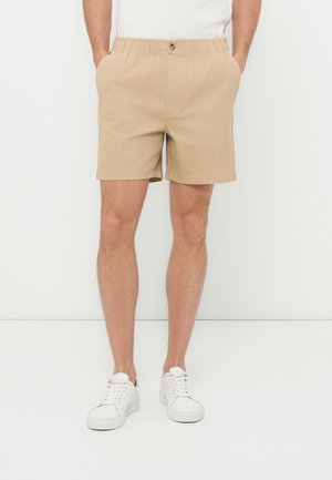 Άνδρας που φοράει μπεζ σορτς με ελαστική μέση και κουμπί και λευκά αθλητικά παπούτσια, στάσιμος με τα χέρια στις τσέπες, μπροστά σε ένα λευκό φόντο.