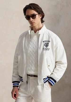 Jeune homme portant des lunettes de soleil écaille de tortue, une veste blanche Wimbledon avec des poignets rayés bleus, un pull en maille torsadée blanche et un short blanc, mains dans les poches.