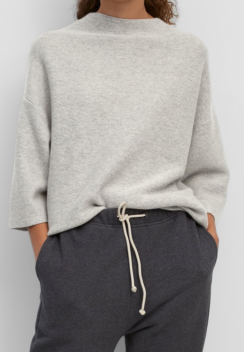 Femme portant un pull en maille texturée gris clair avec des manches aux coudes et un pantalon de survêtement gris foncé avec un cordon blanc noué et les mains dans les poches.