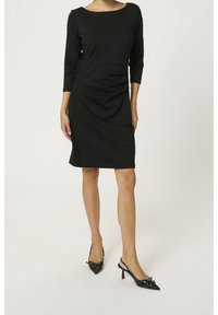 Robe noire à mi-longueur avec manches trois-quarts, présentant un décolleté arrondi et des fronces sur les côtés. Portée avec des escarpins noirs à talon ouvert.